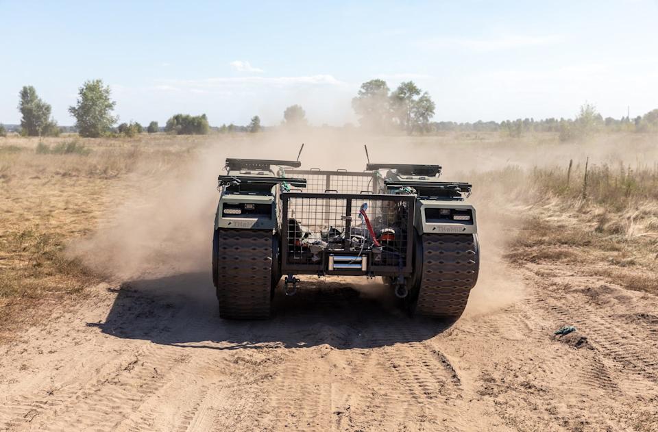 A robot with two sets of tracks and a large wire area for a human to lie drives along a dusty road