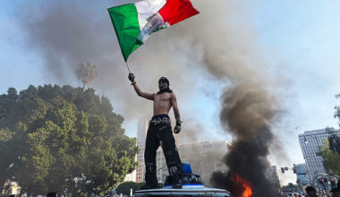 A protester holds a Mexican flag while standing on a vandalized Waymo car during a protest against immigration raids in Los Angeles, California, June 8th 2025