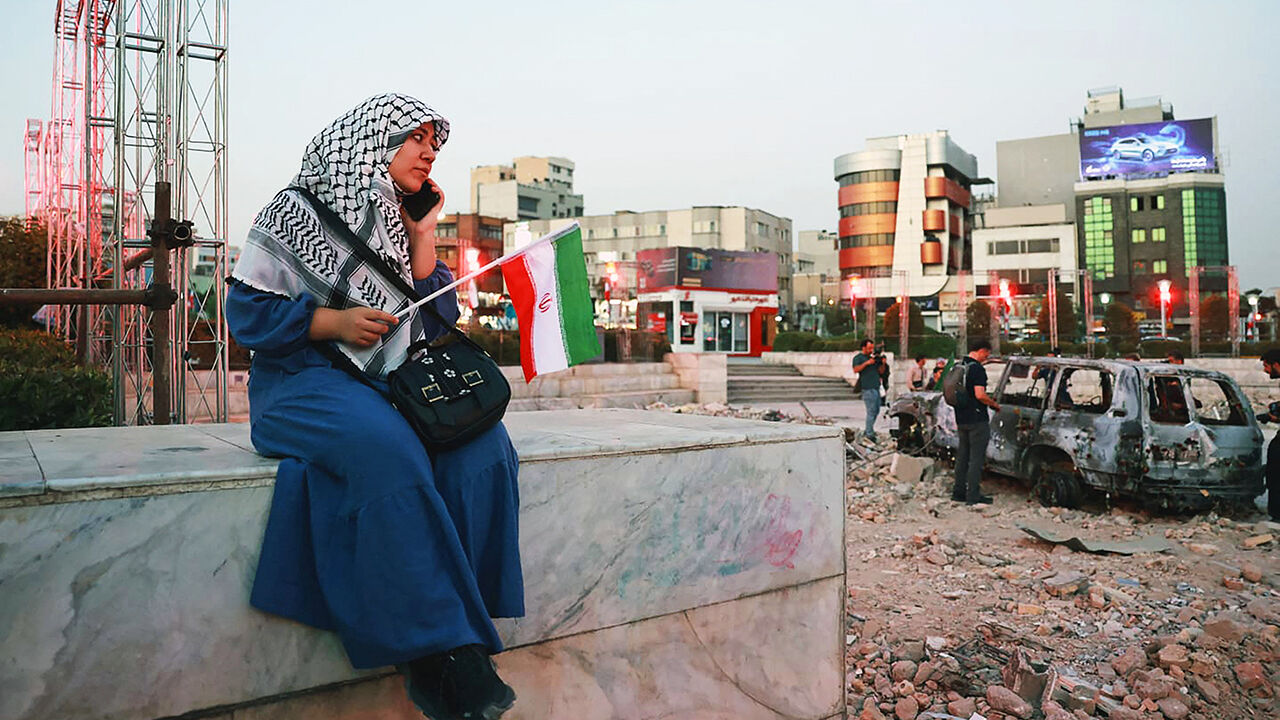 An Iranian woman sits close to a destroyed vehicle as she watches and listens to a traditional band as it performs to show solidarity with the Iranian people at Hafteh-tir Square, in Tehran