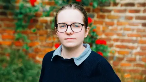 Generation UK A woman with her hair tied back and wearing glasses, looks into the camera. She is wearing a navy jumper and shirt. A brick wall and bushes can be seen in the blurred background behind her.