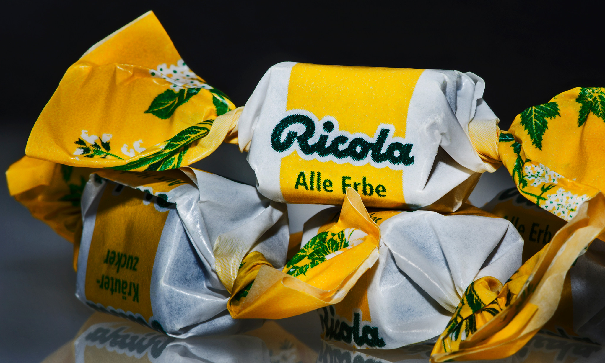 Des bonbons Ricola, emballés en jaune et blanc avec l’inscription ’Ricola Alle Erbe’, quelques cubes jaunes illustrant des plantes.