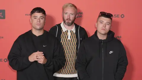 PA Media Kneecap members Moglai Bap, Mo Chara, DJ Provai standing together in front of a red wall. 