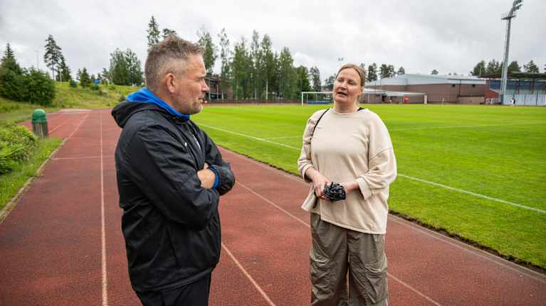 Valmentaja Teemu Eskola ja Diekon Dorren äiti Nina Kuoksa keskustelemassa urheilukentän laidalla pilvisenä päivänä. Taustalla on urheiluhalli ja jalkapallokenttä.