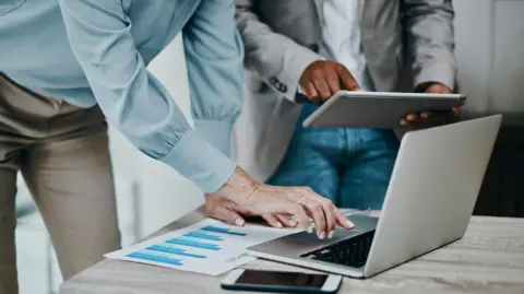 Getty Images Two people whose heads cannot be seen.  One is wearing a blue shirt and beige trouers, the other wears a light-coloured jacket. They are both crouching over laptops. The person nearest the camera is typing on the keyboard. There is a printout of a graph with blue bars on the table alongside a mobile phone.