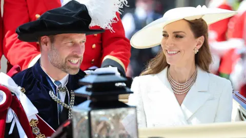 PA Media The Prince and Princess of Wales in a horse carriage. The prince wears ceremonial robes and hat, the princess is in a white jacket and white hat with a pearl necklace.