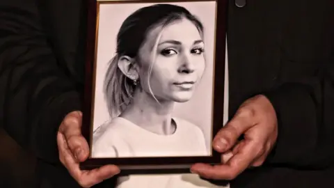 Global Images Ukraine via Getty Images A woman holds a black and white photo of Viktoriia Roshchyna, which shows her looking straight down the barrel of the camera, and her hair tired into a pony tail.