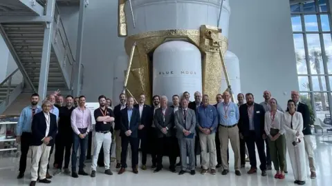Space North East A delegation of space industry representatives standing in front of a Blue Moon lunar lander. The large craft is white with sections painted gold.
