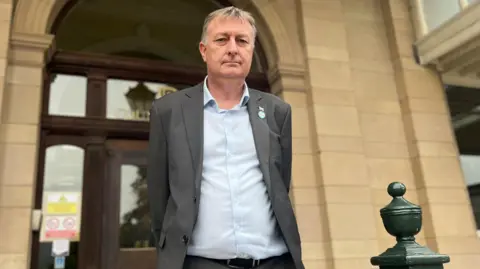 BBC Man with grey hair in grey suit with light blue shirt stood outside a beige stone building with wooden door entrance