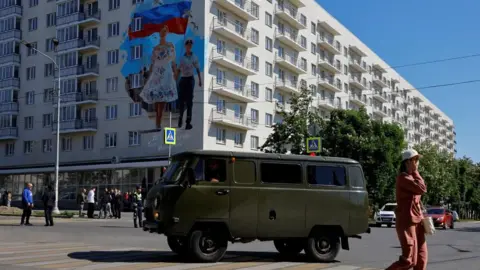REUTERS/Alexander Ermochenko A green van drives through the centre of Mariupol with a Russian mural in the background and a woman wearing a pink jump suit crossing the road