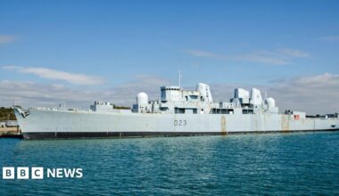An enormous grey warship docked on a quayside on a sunny day. The hull has a couple of rust marks.
