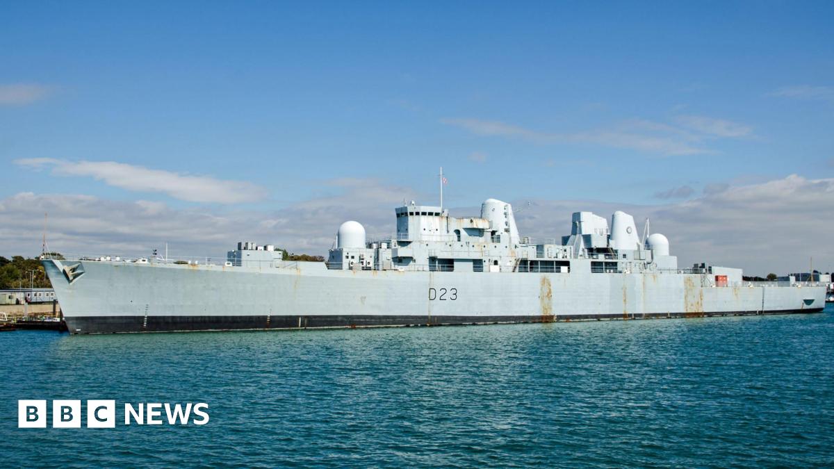 An enormous grey warship docked on a quayside on a sunny day. The hull has a couple of rust marks.