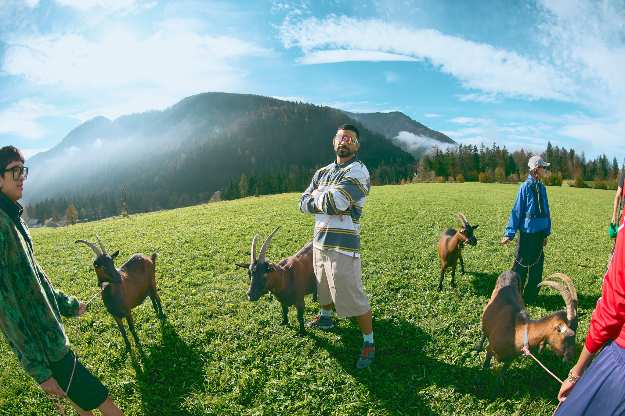 Des personnes entourées de chèvres brunes dans un champ herbeux avec des montagnes en arrière-plan, sous un ciel partiellement couvert. Des personnes entourées de chèvres brunes dans un champ herbeux avec des montagnes en arrière-plan, sous un ciel partiellement couvert.