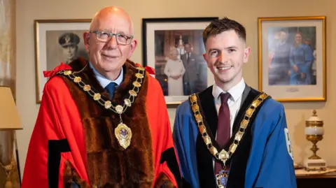 Mid and East Antrim Council Mayor Jackson Minford and Deputy Mayor Tyler Hoey pictured smiling at the camera in an office. Minford is wearing a red, fur-trimmed ceremonial robe over a grey suit, with the gold chain of office around his neck. He has grey-rimmed glasses and no hair. Hoey has short, dark hair and is wearing a blue ceremonial robe and chain over a grey suit with a white shirt and burgundy tie.
