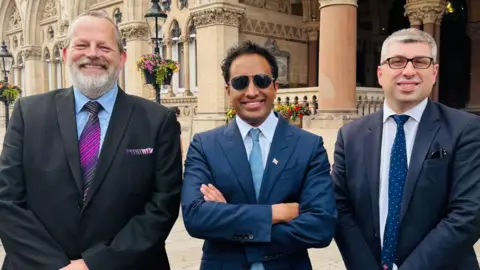 Reform UK James Petter with short grey hair and a beard, Zia Yusuf with short dark hair and dark glasses, and Mark Arnull with short grey hair and glasses all smiling at the camera and wearing suits. The stone pillars of Northampton Guildhall are behind them.