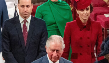 Prince William is wearing a dark suit with a red tie, while Kate Middleton wears a red coat dress and matching hat, and King Charles walks ahead of them in a striped blue suit