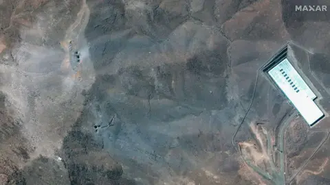Satellite image (c) 2025 Maxar Technologies via Getty Images Maxar Satellite Imagery shows the ridge above the Fordo complex post-strike. Multiple impact craters and a wide grey-blue ash layer are visible across the ridge, suggesting direct hits to the overburden covering the underground facility. 