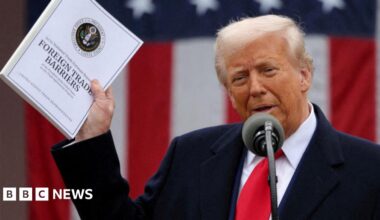Donald Trump holds a document that says 'Foreign Trade Barriers'. He wears a blue suit, white shirt and red tie. There is a mic in front of him and a US flag behind.