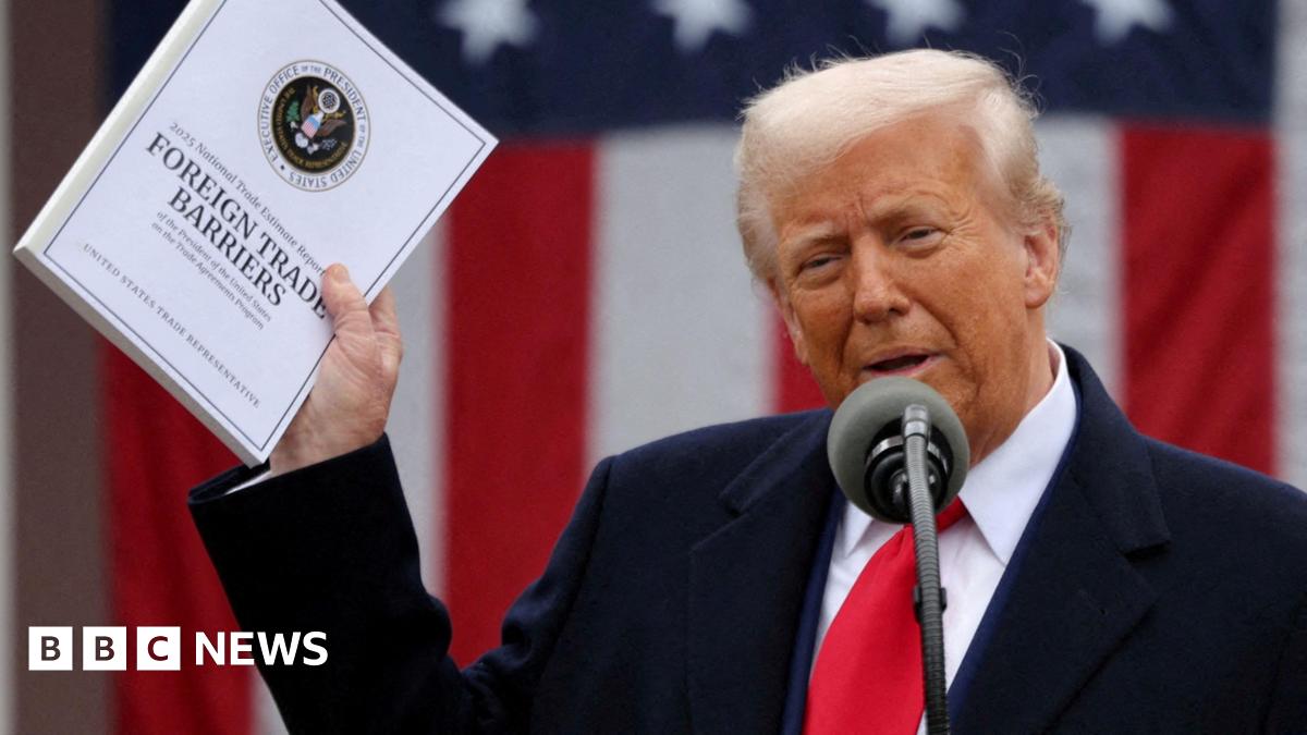 Donald Trump holds a document that says 'Foreign Trade Barriers'. He wears a blue suit, white shirt and red tie. There is a mic in front of him and a US flag behind.