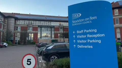 BBC Stockton Council's headquarters Dunedin House. It is a four-storey building mostly made of brown tiles and glass, with solar panels on the roof. A blue sign in the foreground is pointing towards the reception and parking. There are several parked cars and 5mph red and white speed restriction sign.