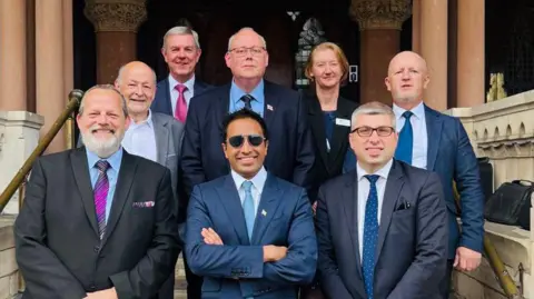 Reform UK A group of seven men and one woman, formally dressed, standing on steps in Northampton's pillared Guildhall.  Front centre is Zia Yusuf, with short dark hair and glasses, wearing a blue jacket and tie with his arms folded.