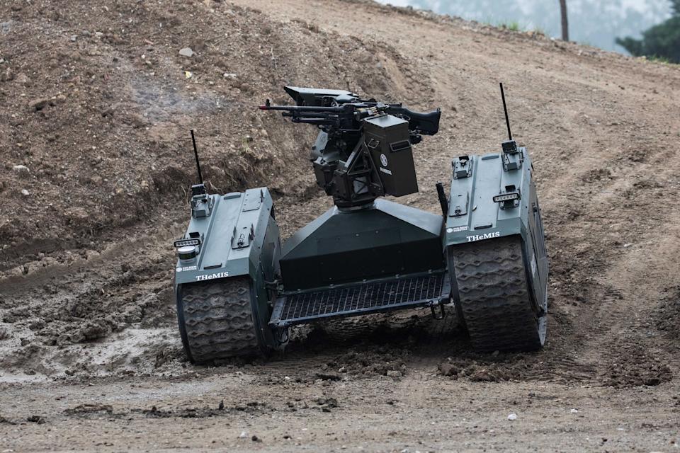 A dark green robot with tracks and a gun on top drives on a muddy hill