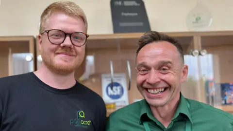 BBC Two men stand side by side, both smiling. On the left, Aran has ginger hair and a ginger beard. He is also wearing a pair of rounded burgundy glasses. Grant, an older man, has brown hair and wears a green shirt. Behind them, a blurred view of a cabinet with a display of company awards.
