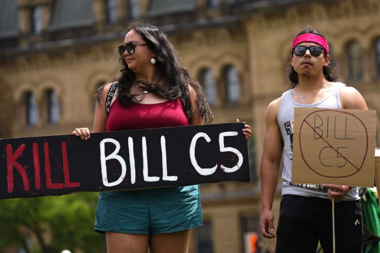 Two protesters holding signs—one of which reads "Kill bill C5"—against the blurred backdrop of the Parliament Buildings in Ottawa.