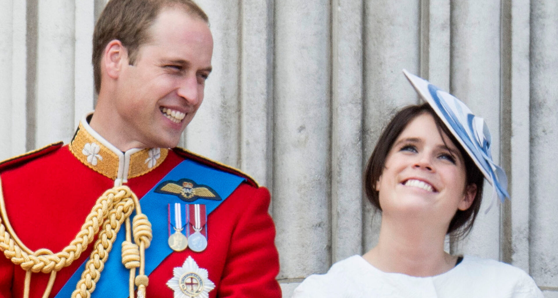 Prince William laughs while wearing red military uniform as he stands next to Princess Eugenie, who is wearing a white dress and a blue and white hat on the Buckingham Palace balcony