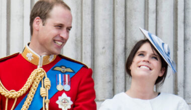 Prince William laughs while wearing red military uniform as he stands next to Princess Eugenie, who is wearing a white dress and a blue and white hat on the Buckingham Palace balcony