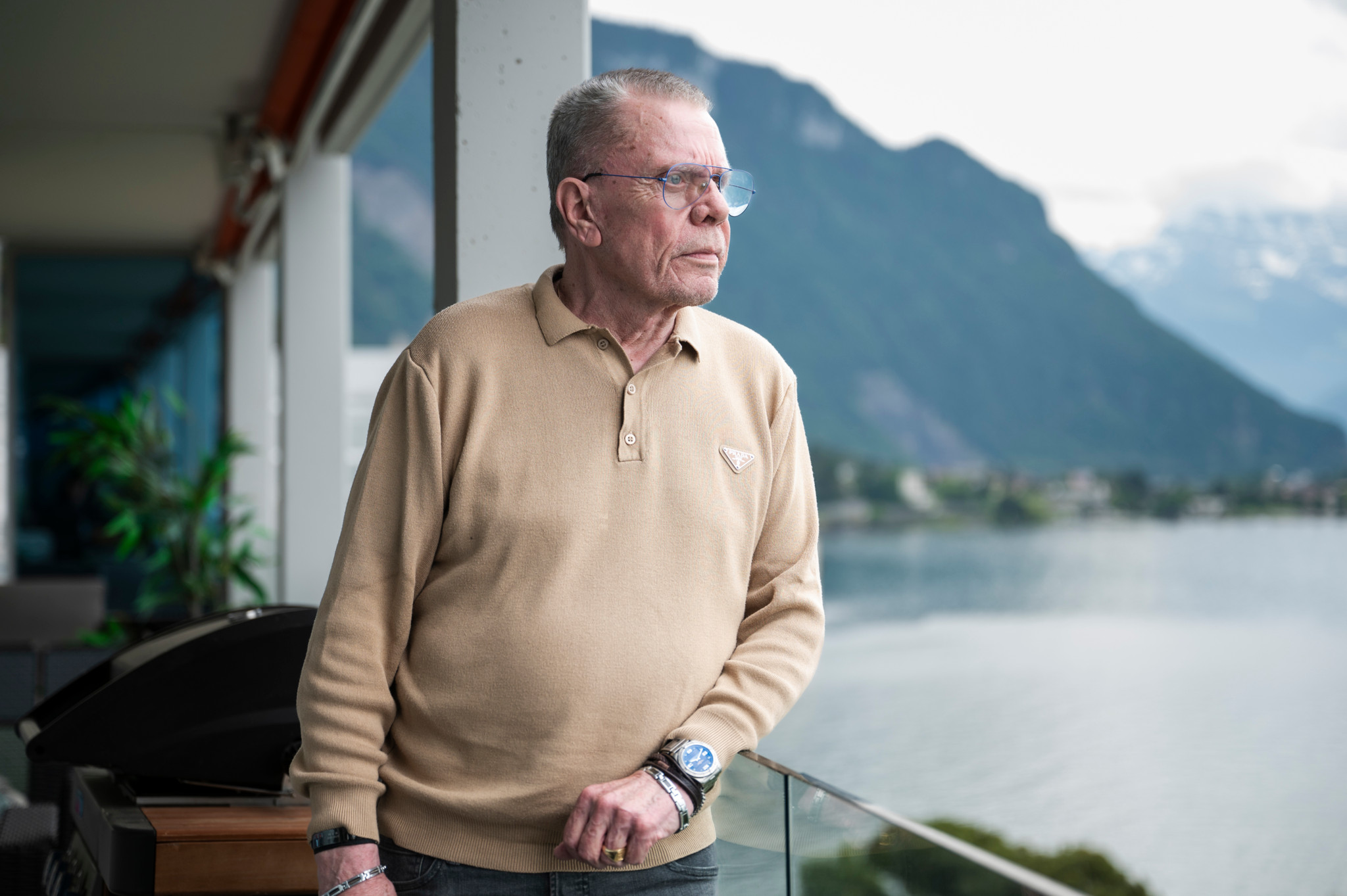 Martin Jeanneret, portant un pull beige, se tient sur un balcon à Veytaux, avec une vue sur le lac et les montagnes en arrière-plan.