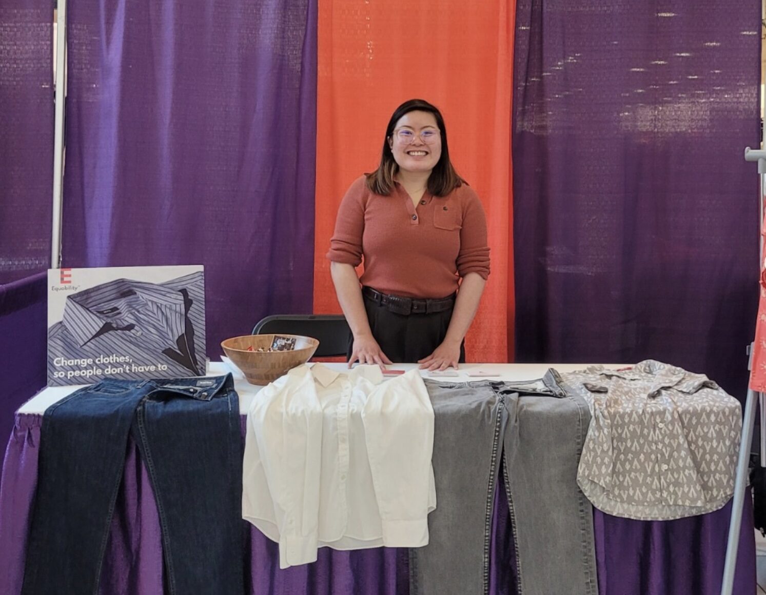 A woman stands behind a booth with a sign reading Equability LLC, displaying clothing items including jeans, shirts, and a patterned top. A small sign and basket are on the table.