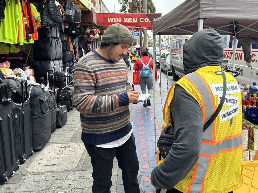 A man in a striped sweater talks to a street vendor in a yellow vest on a busy sidewalk lined with shops and merchandise, capturing everyday life as seen by Mission Local.
