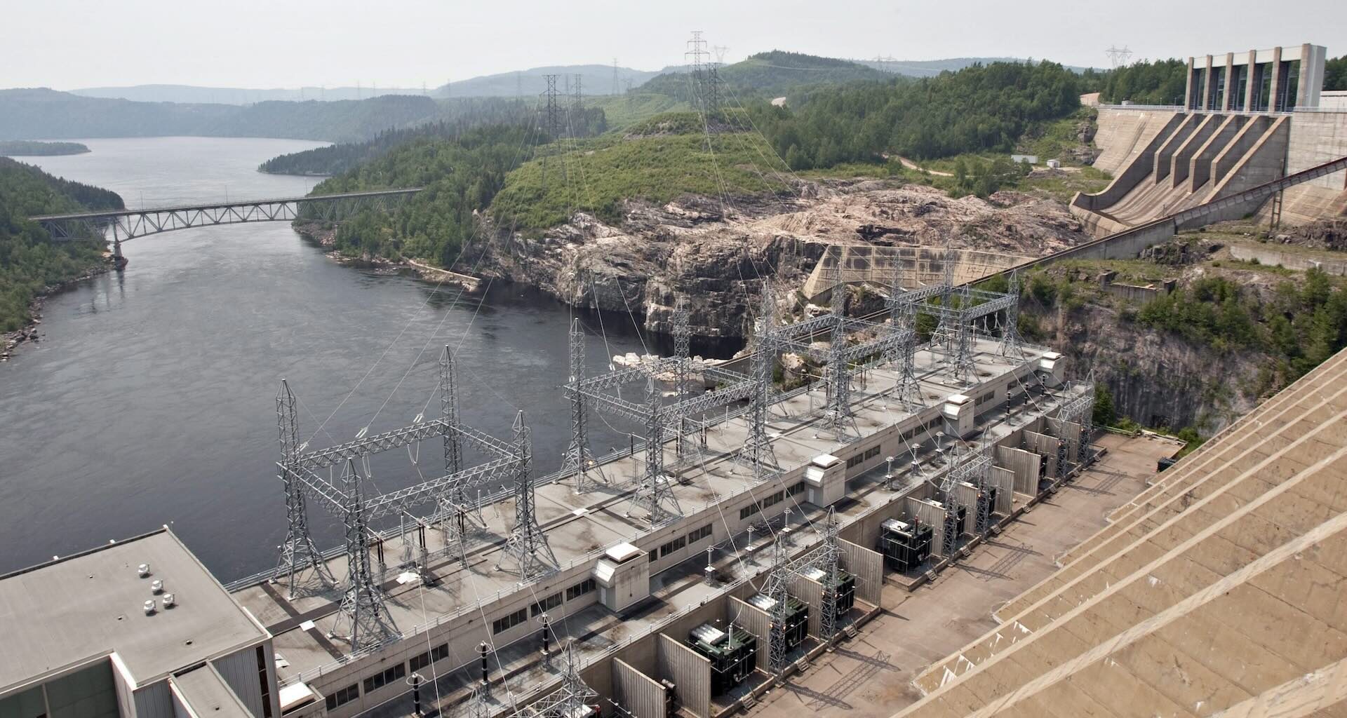 A large hydroelectric dam with transmission lines spans a river, with forested hills, a bridge, and industrial structures visible in the background.
