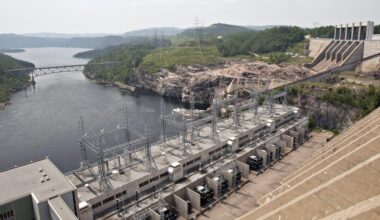 A large hydroelectric dam with transmission lines spans a river, with forested hills, a bridge, and industrial structures visible in the background.