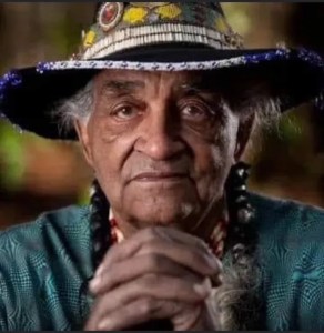 An elderly Native American man looks forward with his chin resting on clasped hands. He has braids and is wearing a wide-brimmed hat with embroidery on it.