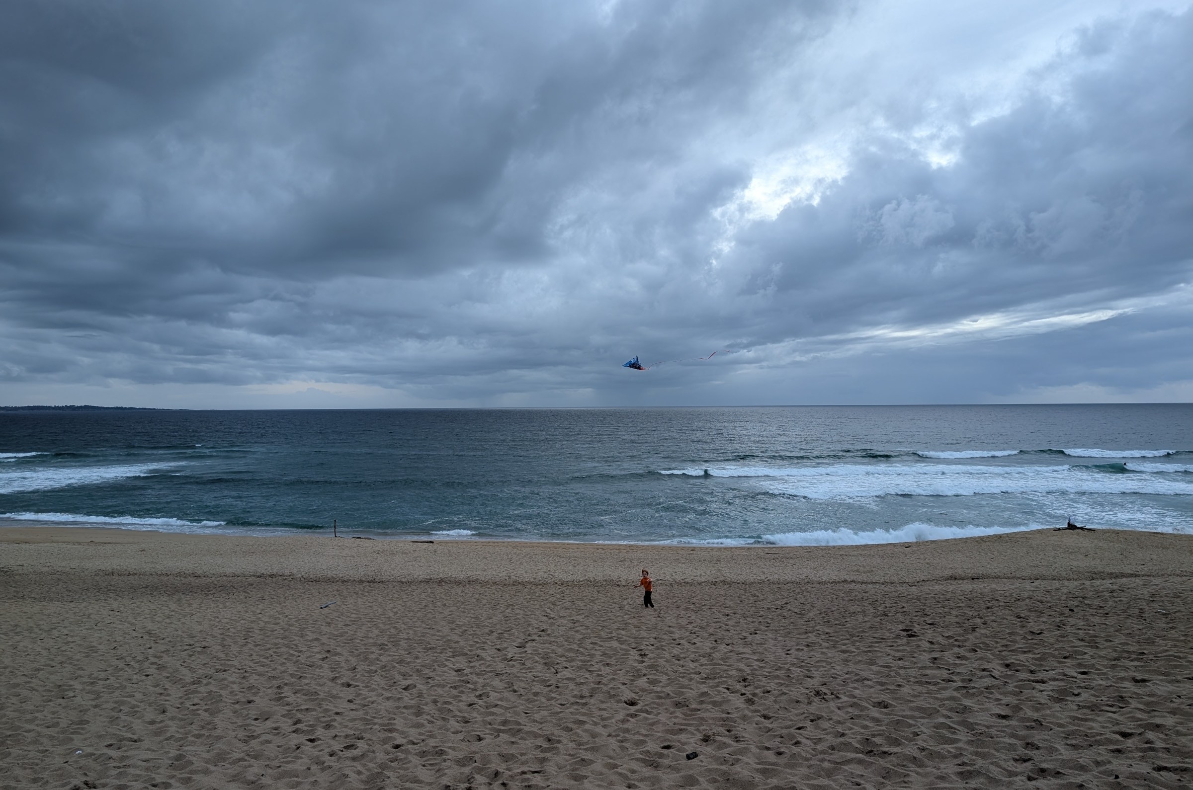 A child is a small figure on a cloudy beach, flying a kite.