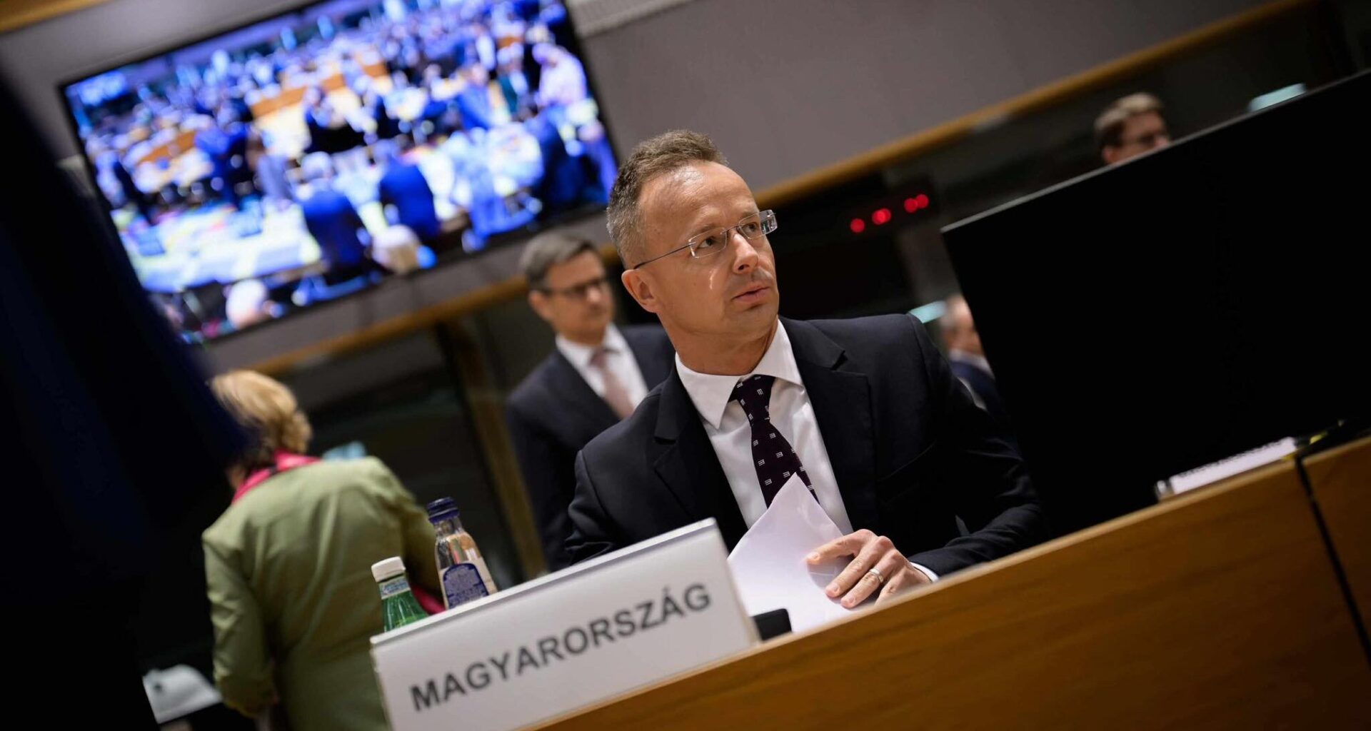 Hungarian foreign minister Peter Szijjartó seated, backdrop of television showing other Council of the EU members getting seated