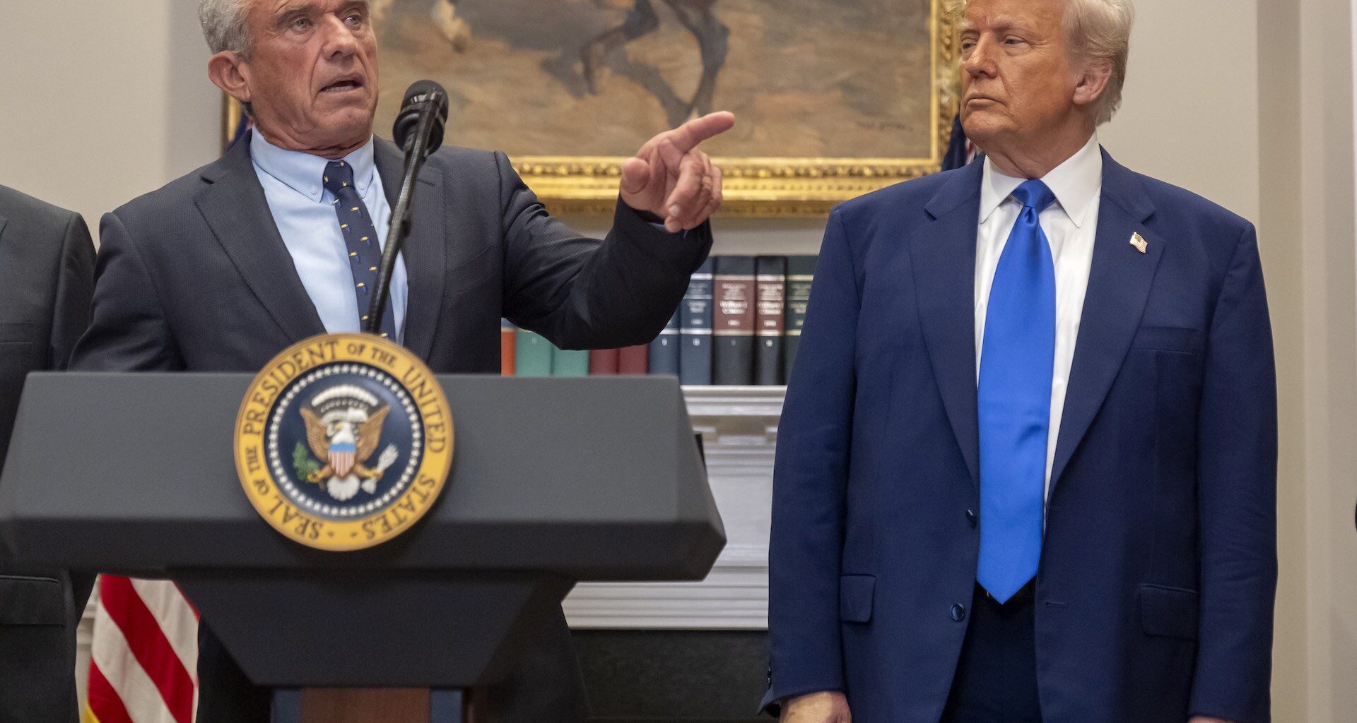 A lower-angle shot of Robert F. Kennedy Jr. behind a lectern with the U.S. presidential seal on the front. He is pointing off-camera. Donald Trump is standing to Kennedy's left.