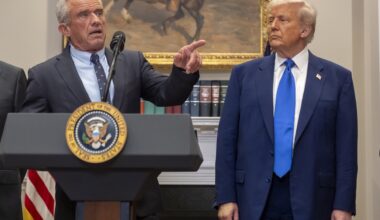 A lower-angle shot of Robert F. Kennedy Jr. behind a lectern with the U.S. presidential seal on the front. He is pointing off-camera. Donald Trump is standing to Kennedy's left.