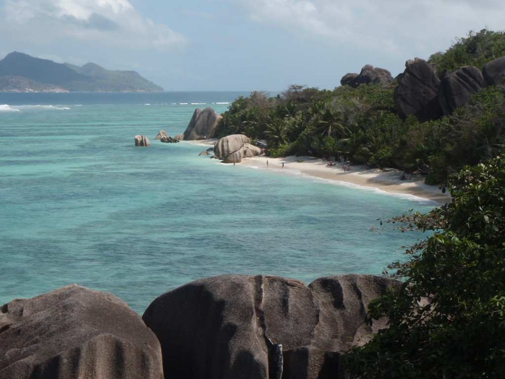A landscape view of the coastline along an island in the Seychelles. The turquoise blue water hubs the sandy beach. Beyond the beach are limestone rocks, palm trees and other tropical foliage.