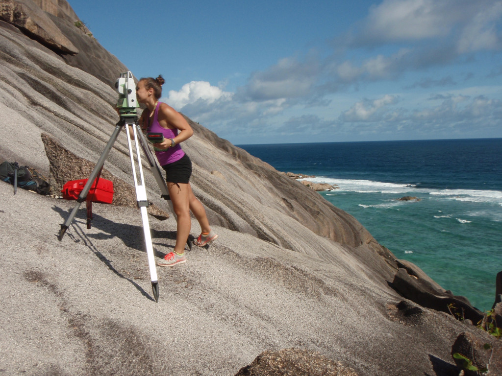 Vyverberg stands on a rocky outcrop and peers through a total station to survey the elevation of fossil corals on La Digue, Seychelles. The modern reef is visible in the background.