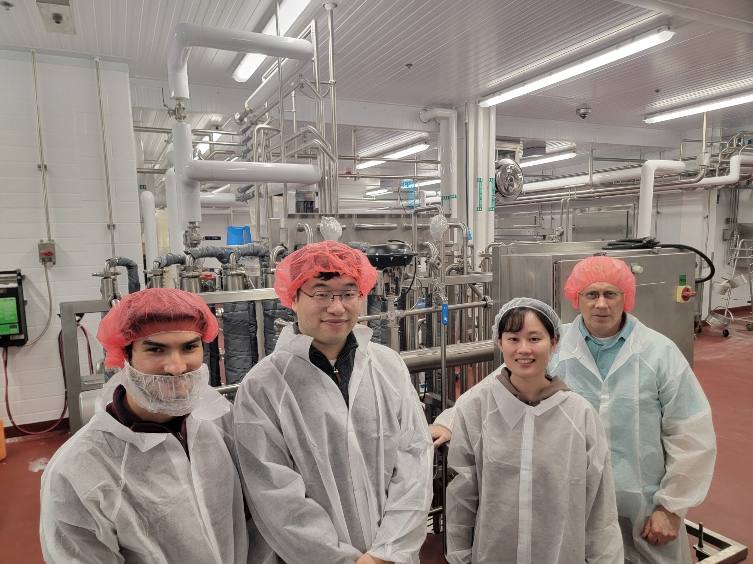 Four people wearing lab coats, hairnets, and beard covers stand in a food processing or laboratory facility with industrial equipment in the background.
