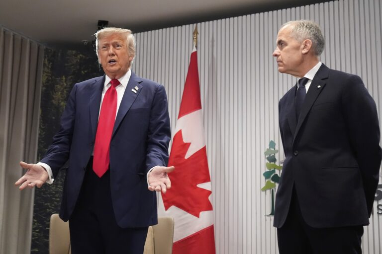 A shot of Donald Trump and Mark Carney in front of a Canadian flag on a stand, and a backdrop bearing the 2025 G7 summit logo. Trump is speaking with his hands slightly spread, and Carney is looking at him intently.