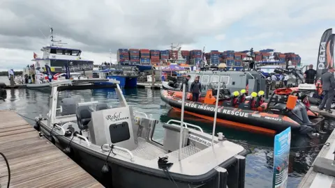 BBC A silver open deck power boat is tied up at a pontoon with a lifeboat and a large container ship in the background