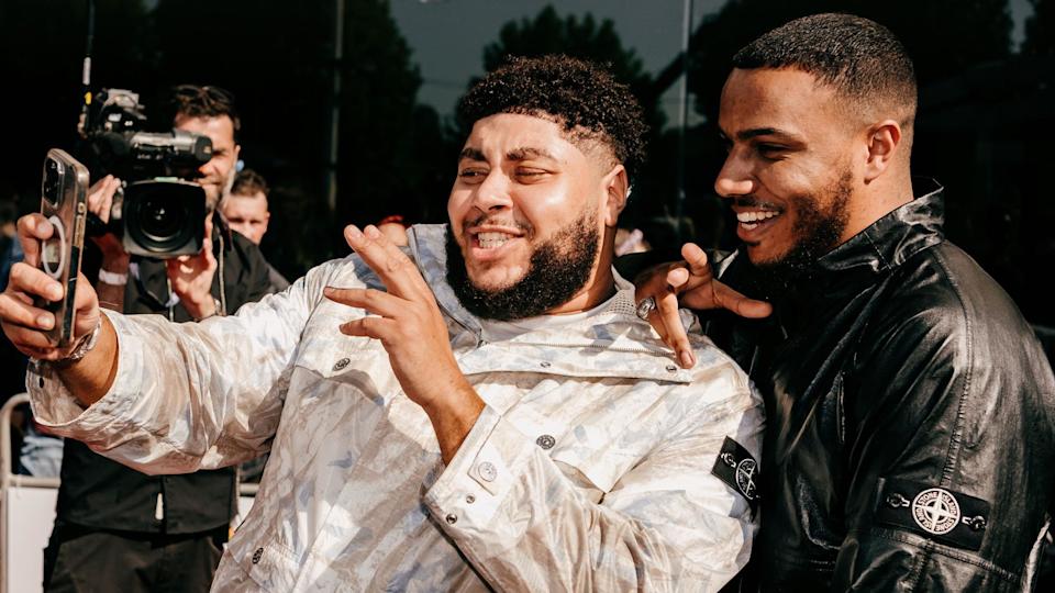 AJ Tracey and Big Zuu taking a selfie on a red carpet. AJ (right) smiles over Big Zuu's shoulder as he holds up his phone. AJ wears a black jacket while Zuu is in a white coat. It's sunny outside and members of the media can be seen behind them. 