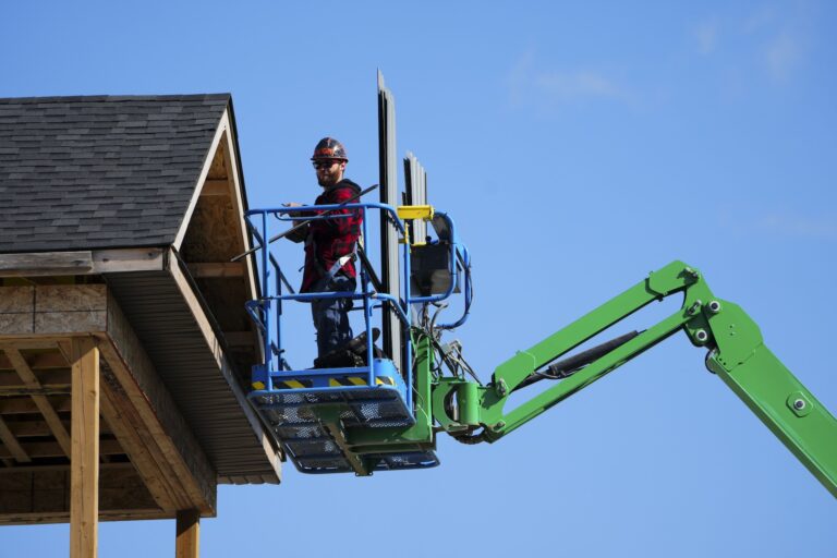 A construction worker stands on a green boom lift, working on the exterior of a building under a clear blue sky.