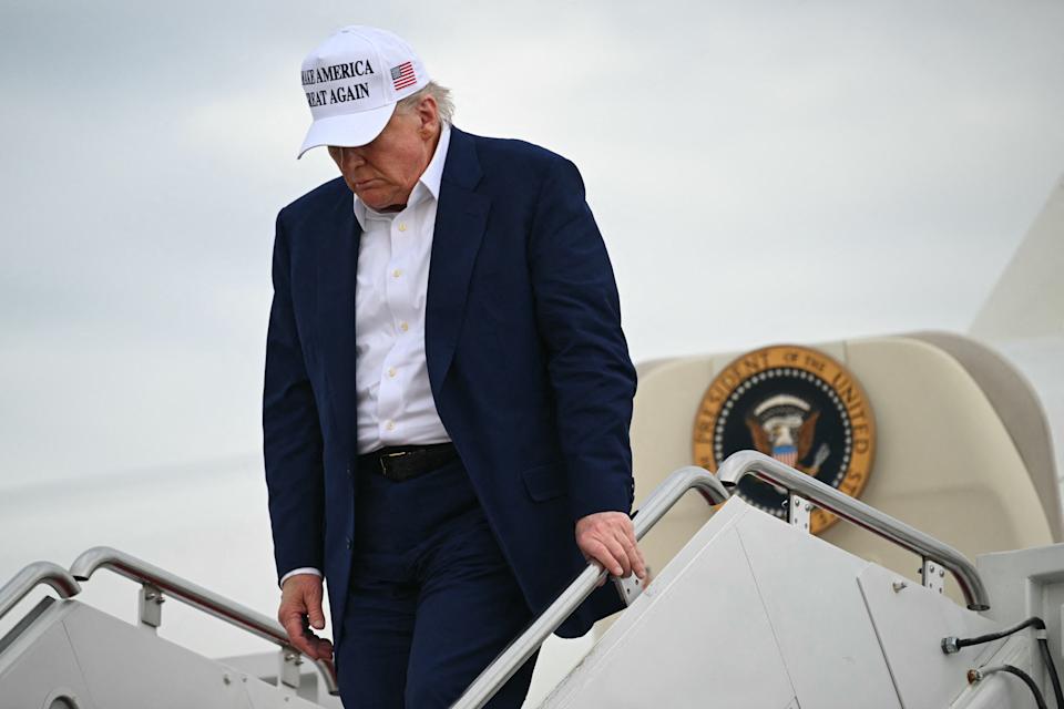 Person in a suit and hat descends airplane steps, near a large emblem