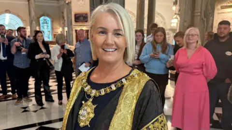 BBC Belfast Lord Mayor Tracy Kelly wearing her ceremonial robe and gold chain of office in Belfast City Hall. She has short, bobbed blonde hair and is smiling at the camera. A number of people are standing behind her watching inside the hall.