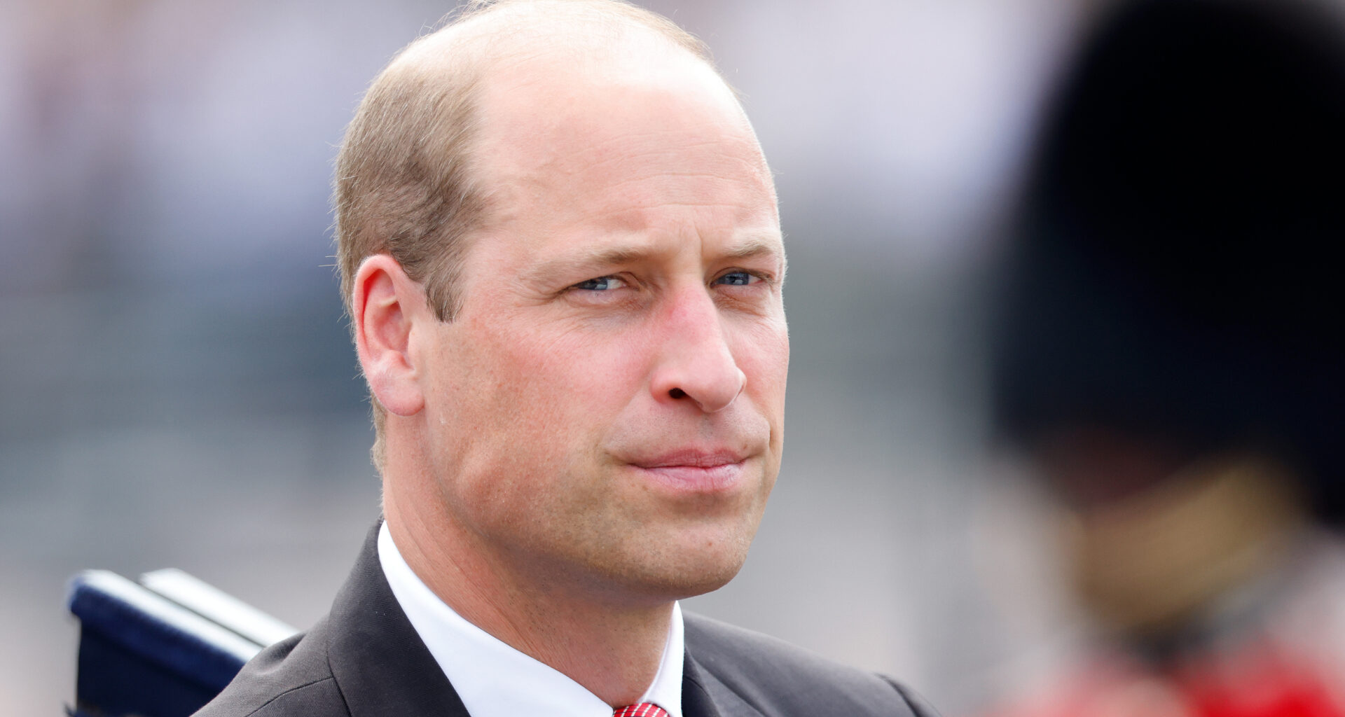 Prince William wears a black suit with a red tie and looks serious