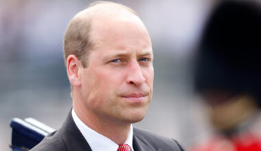Prince William wears a black suit with a red tie and looks serious